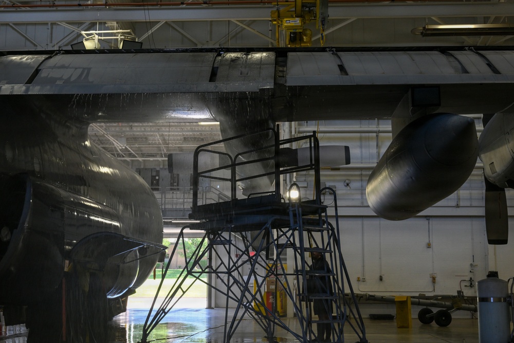 The image shows the underside of a large military aircraft, likely a C-130 or similar transport aircraft, inside a maintenance hangar. A maintenance scaffold/work platform is positioned beneath the aircraft's fuselage and engine nacelles, with a work light illuminating the area. The aircraft appears wet, suggesting a recent wash or cleaning procedure.