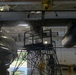 The image shows the underside of a large military aircraft, likely a C-130 or similar transport aircraft, inside a maintenance hangar. A maintenance scaffold/work platform is positioned beneath the aircraft's fuselage and engine nacelles, with a work light illuminating the area. The aircraft appears wet, suggesting a recent wash or cleaning procedure.