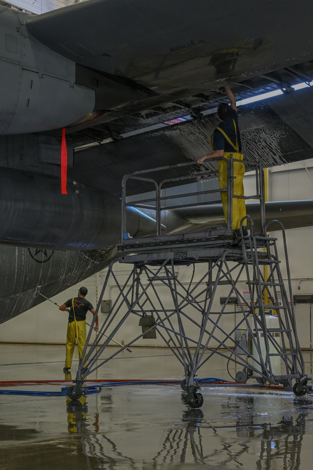Two maintenance workers in yellow waterproof overalls are washing the underside of a large military aircraft inside a hangar. One worker stands on a wheeled maintenance platform (marked 'B509') scrubbing the aircraft's belly, while a second worker on the floor uses a long-handled brush. The hangar floor is wet with soapy water and hoses are visible running across the floor.