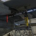 Two maintenance workers in yellow waterproof overalls are washing the underside of a large military aircraft inside a hangar. One worker stands on a wheeled maintenance platform (marked 'B509') scrubbing the aircraft's belly, while a second worker on the floor uses a long-handled brush. The hangar floor is wet with soapy water and hoses are visible running across the floor.