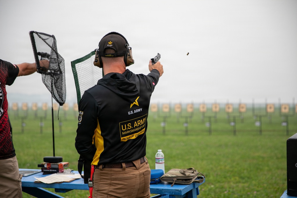 A U.S. Army Marksmanship Unit competitor is actively firing a pistol at an outdoor shooting range, with a spent casing visibly ejecting from the firearm. The shooter is wearing official U.S. Army Marksmanship Unit gear including a black and gold jacket, matching cap, and hearing protection. Multiple paper targets are visible downrange in a row across a grassy field.