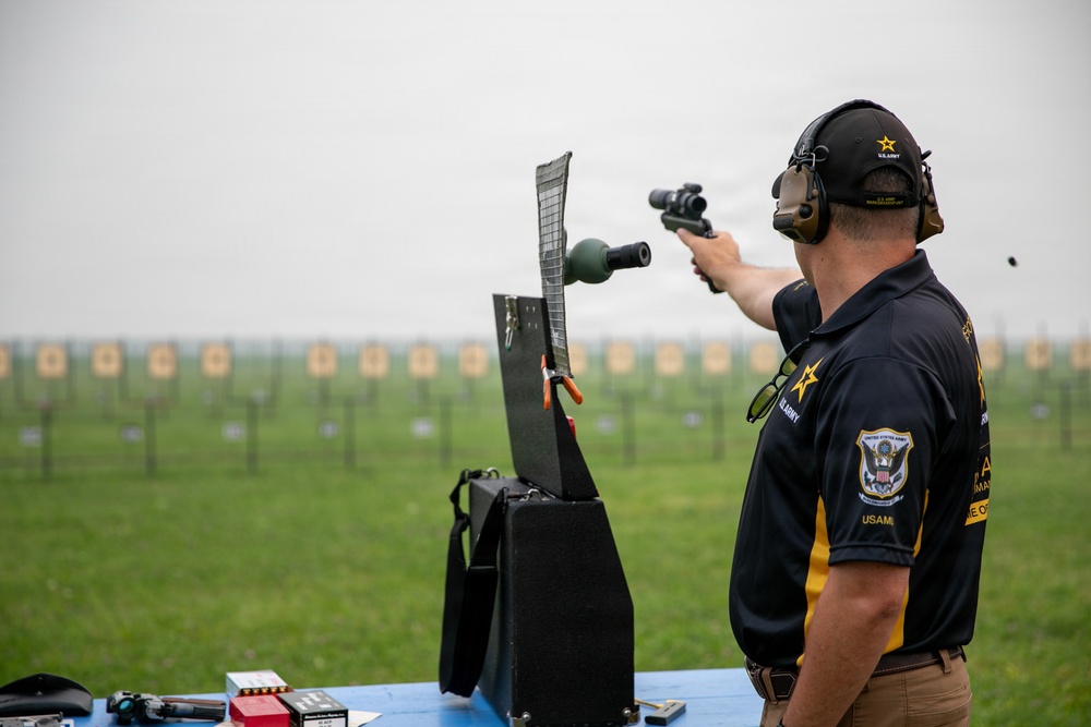 A U.S. Army Marksmanship Unit (USAMU) competitive shooter is firing a precision pistol at an outdoor shooting range. The shooter is equipped with hearing protection, a scoped pistol, and is wearing official USAMU team apparel. A row of distant paper targets is visible downrange across a large grass field.
