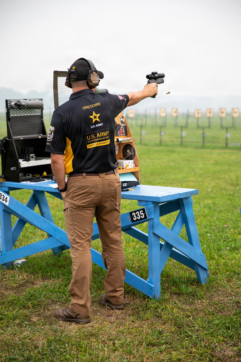 A competitive shooter identified as 'GREGOIRE' from the U.S. Army Marksmanship Unit is actively firing a pistol at an outdoor shooting range. A spent brass casing is visibly ejecting from the firearm mid-shot. The shooter stands at station 335, wearing full competition attire including hearing protection and a U.S. Army Marksmanship Unit uniform.