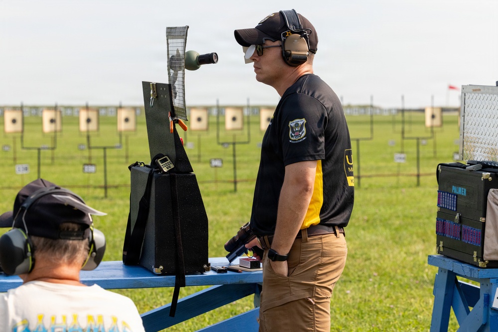 A competitive pistol shooter wearing hearing protection, sunglasses, and a black military-style team shirt stands at a shooting bench on an outdoor range. He is holding a pistol and appears to be preparing for or between shooting stages. Multiple paper targets are visible downrange on a well-maintained grass range.