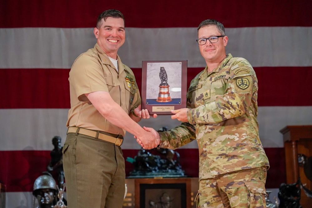Two military personnel from different branches are posing together during an award ceremony, jointly holding a trophy plaque while shaking hands. The individual on the left is wearing a U.S. Navy service uniform (tan/khaki), while the individual on the right is wearing U.S. Army OCP (Operational Camouflage Pattern) uniform. An American flag serves as the backdrop, and additional trophies are visible in the background.