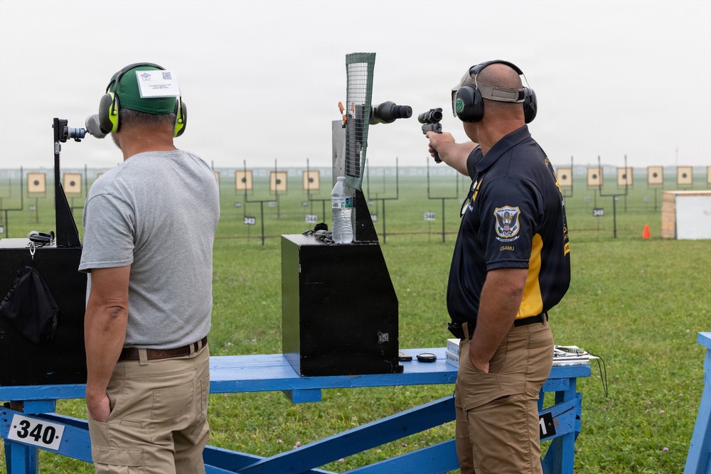 Two men are at an outdoor shooting range, with one actively aiming a pistol downrange while the other observes. The man shooting wears a dark navy polo shirt with what appears to be a U.S. Army Marksmanship Unit (USAMU) emblem, suggesting a competitive or official military shooting event. Multiple paper targets are visible in the background across a large grass range.