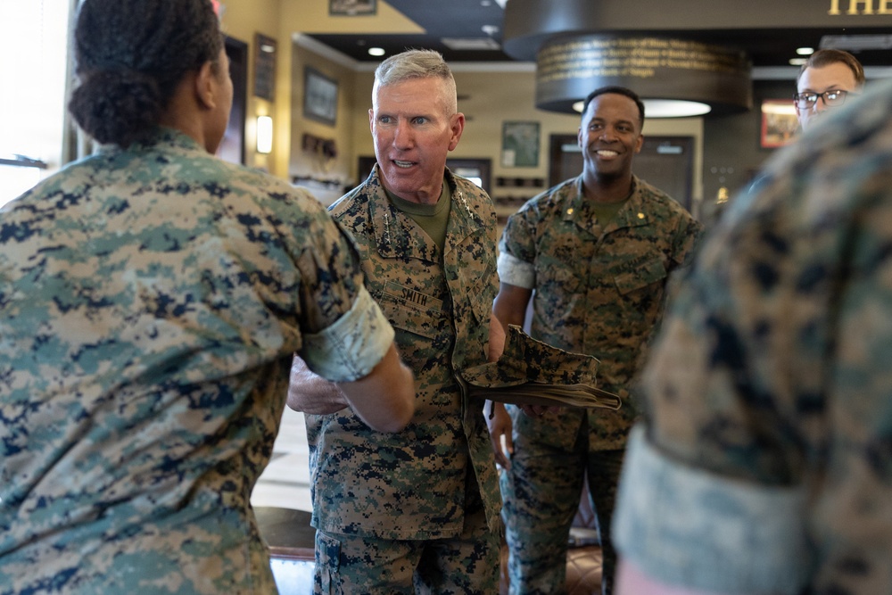 Several U.S. Marine Corps personnel in MARPAT digital camouflage uniforms are gathered in what appears to be an indoor military facility. A central figure with the name tape 'SMITH' is shaking hands with another service member while holding a folder. The atmosphere appears cordial and professional, suggesting an official visit or greeting ceremony.