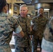 Several U.S. Marine Corps personnel in MARPAT digital camouflage uniforms are gathered in what appears to be an indoor military facility. A central figure with the name tape 'SMITH' is shaking hands with another service member while holding a folder. The atmosphere appears cordial and professional, suggesting an official visit or greeting ceremony.