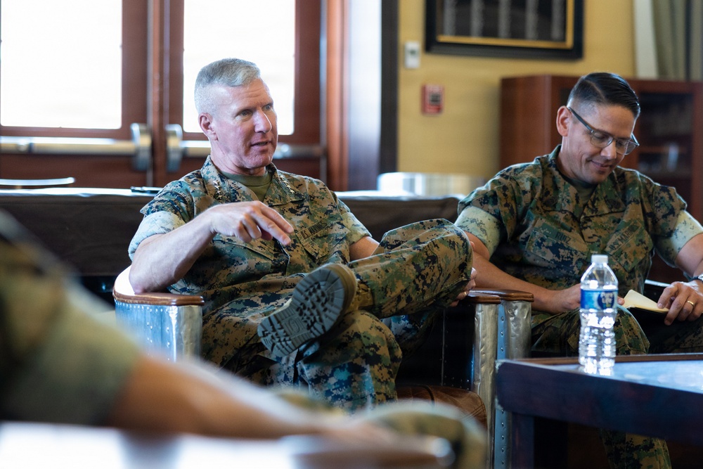 Two military personnel in U.S. Marine Corps MARPAT camouflage uniforms are seated in a formal meeting or briefing setting. The individual on the left appears to be speaking and gesturing, while the one on the right is listening and taking notes. The setting appears to be an office or conference room with wooden furnishings.
