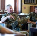 Two military personnel in U.S. Marine Corps MARPAT camouflage uniforms are seated in a formal meeting or briefing setting. The individual on the left appears to be speaking and gesturing, while the one on the right is listening and taking notes. The setting appears to be an office or conference room with wooden furnishings.