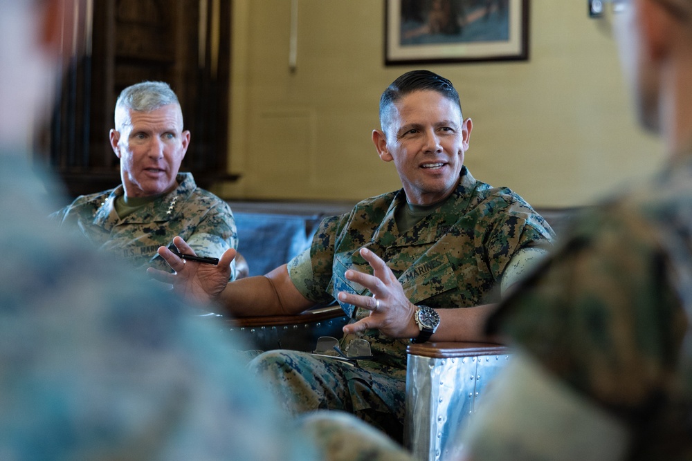 Two U.S. Marines in MARPAT camouflage uniforms are seated and engaged in what appears to be a formal discussion or briefing in an indoor setting. The central figure is actively speaking and gesturing with his hands, while a second Marine in the background also appears to be participating in the conversation. Additional uniformed personnel are partially visible in the foreground, suggesting a group meeting or leadership discussion.