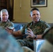 Two U.S. Marines in MARPAT camouflage uniforms are seated and engaged in what appears to be a formal discussion or briefing in an indoor setting. The central figure is actively speaking and gesturing with his hands, while a second Marine in the background also appears to be participating in the conversation. Additional uniformed personnel are partially visible in the foreground, suggesting a group meeting or leadership discussion.