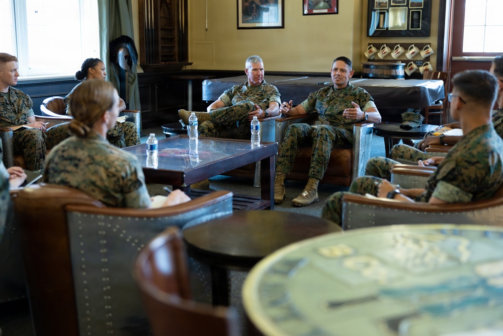 A group of U.S. Marines in woodland MARPAT camouflage uniforms are seated in a lounge-style room engaged in what appears to be an informal briefing or discussion. Two senior Marines seated on a central sofa appear to be leading the conversation, with several other Marines seated around them in leather chairs. The setting suggests a command or officer's club environment with framed military memorabilia on the walls.