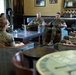 A group of U.S. Marines in woodland MARPAT camouflage uniforms are seated in a lounge-style room engaged in what appears to be an informal briefing or discussion. Two senior Marines seated on a central sofa appear to be leading the conversation, with several other Marines seated around them in leather chairs. The setting suggests a command or officer's club environment with framed military memorabilia on the walls.
