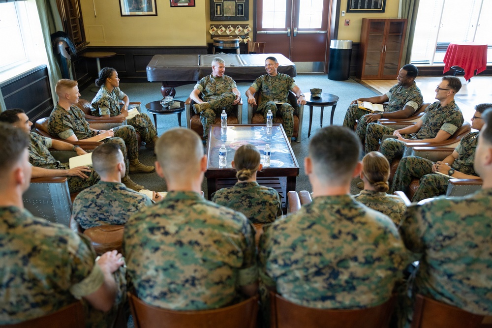 A group of military personnel in MARPAT (Marine digital camouflage) uniforms are seated in a circle in what appears to be a lounge or meeting room. Two senior personnel are seated centrally, appearing to lead or facilitate a discussion with the group. The setting suggests an informal briefing, mentorship session, or leadership engagement.