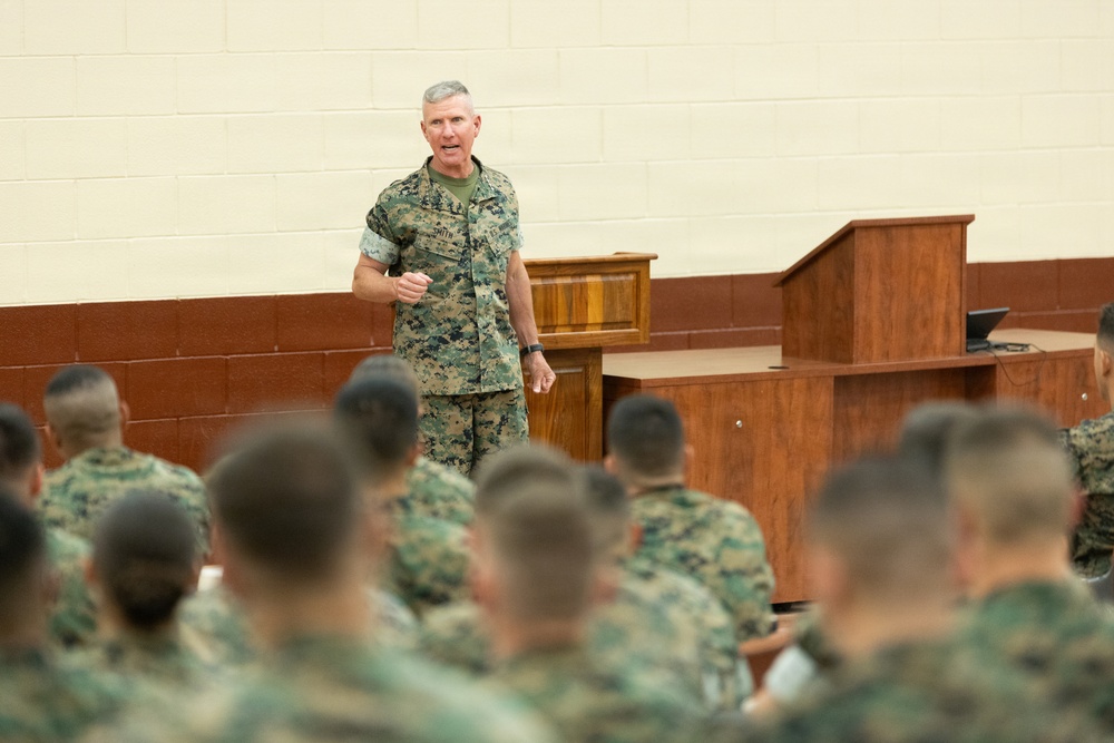 A senior military officer in U.S. Marine Corps MARPAT camouflage uniform is addressing a large group of seated Marines in what appears to be a briefing hall or auditorium. The speaker stands at the front near a wooden podium, gesturing with his hand as he speaks. The audience Marines are seated with their backs to the camera, listening attentively.