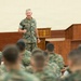 A senior military officer in U.S. Marine Corps MARPAT camouflage uniform is addressing a large group of seated Marines in what appears to be a briefing hall or auditorium. The speaker stands at the front near a wooden podium, gesturing with his hand as he speaks. The audience Marines are seated with their backs to the camera, listening attentively.