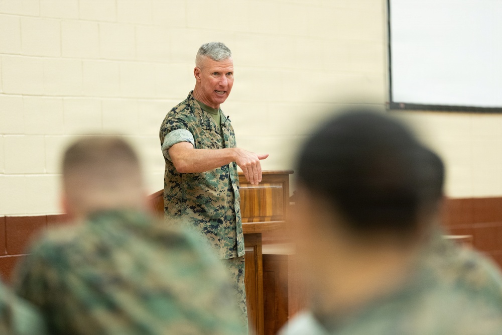 A senior military officer in U.S. Marine Corps MARPAT camouflage uniform is addressing an audience from behind a wooden podium. The speaker appears to be delivering a briefing or lecture to seated personnel, also in camouflage uniforms, visible in the foreground out of focus. The setting appears to be a military briefing room or auditorium with a projection screen visible in the background.
