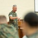 A senior military officer in U.S. Marine Corps MARPAT camouflage uniform is addressing an audience from behind a wooden podium. The speaker appears to be delivering a briefing or lecture to seated personnel, also in camouflage uniforms, visible in the foreground out of focus. The setting appears to be a military briefing room or auditorium with a projection screen visible in the background.