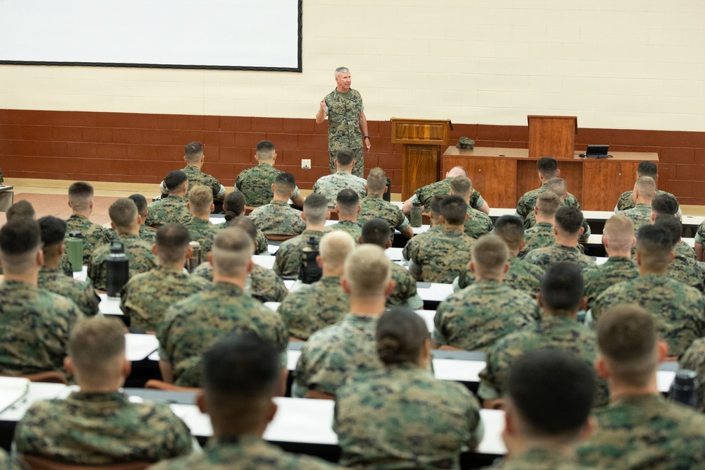 A senior military officer in camouflage uniform addresses a large group of uniformed personnel in an auditorium or lecture hall setting. The audience, all wearing matching woodland camouflage uniforms consistent with U.S. Marine Corps MARPAT pattern, are seated in tiered rows facing the speaker. The setting appears to be a formal military briefing or training session at a military installation.