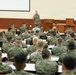 A senior military officer in camouflage uniform addresses a large group of uniformed personnel in an auditorium or lecture hall setting. The audience, all wearing matching woodland camouflage uniforms consistent with U.S. Marine Corps MARPAT pattern, are seated in tiered rows facing the speaker. The setting appears to be a formal military briefing or training session at a military installation.
