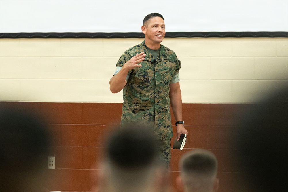 A military personnel wearing USMC MARPAT digital camouflage uniform is addressing an audience in what appears to be a briefing room or gymnasium. The speaker is gesturing with one hand while holding an object in the other, suggesting an active presentation or address. An audience of uniformed individuals is visible in the foreground, slightly out of focus.