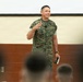 A military personnel wearing USMC MARPAT digital camouflage uniform is addressing an audience in what appears to be a briefing room or gymnasium. The speaker is gesturing with one hand while holding an object in the other, suggesting an active presentation or address. An audience of uniformed individuals is visible in the foreground, slightly out of focus.