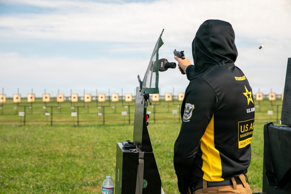 A competitive shooter affiliated with the U.S. Army Marksmanship Unit is firing a pistol at an outdoor shooting range. A spent brass casing is visibly ejecting from the firearm mid-shot. The range features numerous paper targets arranged in rows across a grassy field.