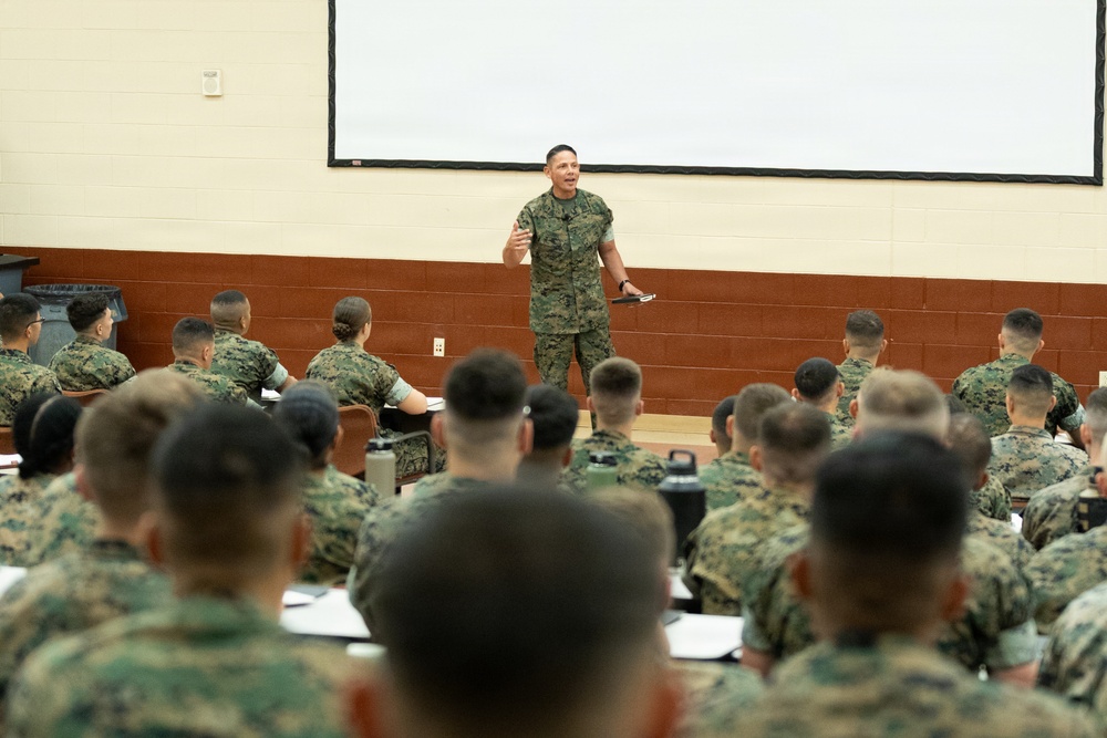 A military briefing or training session is taking place in an auditorium-style classroom. One uniformed service member stands at the front, addressing a large group of seated personnel, all wearing matching camouflage uniforms consistent with U.S. Marine Corps MARPAT pattern. A large projection screen is visible behind the speaker.