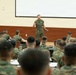 A military briefing or training session is taking place in an auditorium-style classroom. One uniformed service member stands at the front, addressing a large group of seated personnel, all wearing matching camouflage uniforms consistent with U.S. Marine Corps MARPAT pattern. A large projection screen is visible behind the speaker.