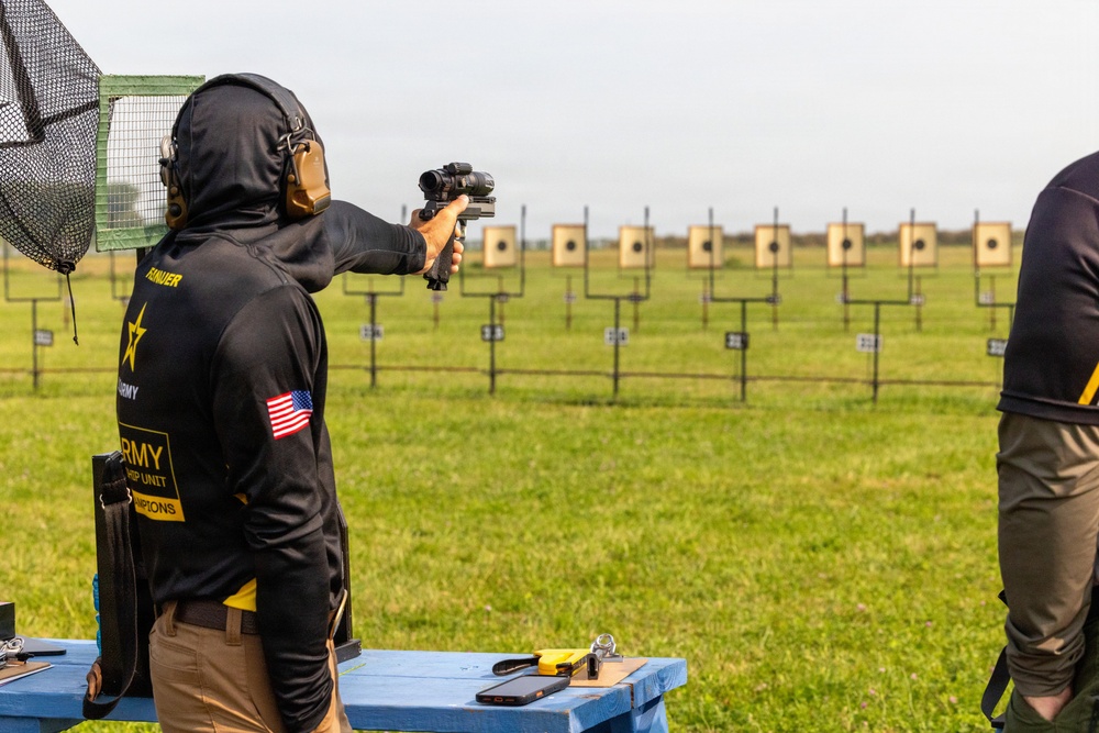 A competitive shooter wearing U.S. Army Marksmanship Unit (AMU) gear is aiming a pistol equipped with an optic sight at a row of paper targets on an outdoor shooting range. The shooter is wearing hearing protection, a black hoodie with an American flag patch, and is positioned at a shooting bench. Multiple square paper targets are visible downrange across a grassy field.