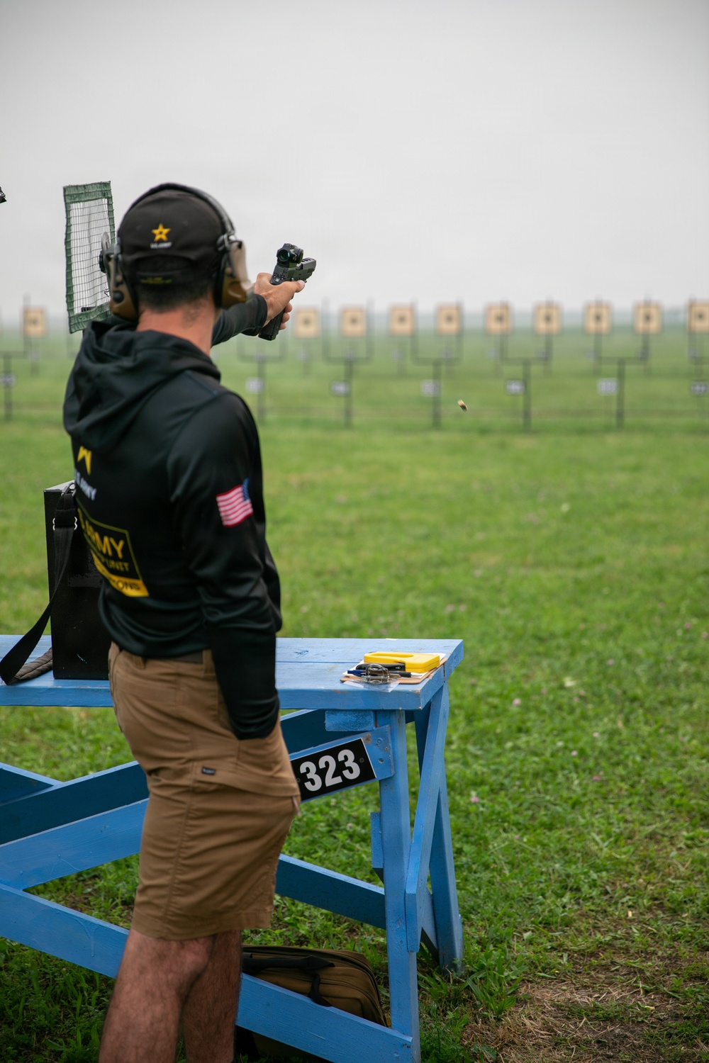 A competitive shooter wearing U.S. Army Marksmanship Unit gear is firing a pistol at an outdoor shooting range. The shooter is positioned at station number 323, with a blue wooden bench beside them holding equipment. A row of paper targets is visible downrange in a misty, overcast outdoor setting.