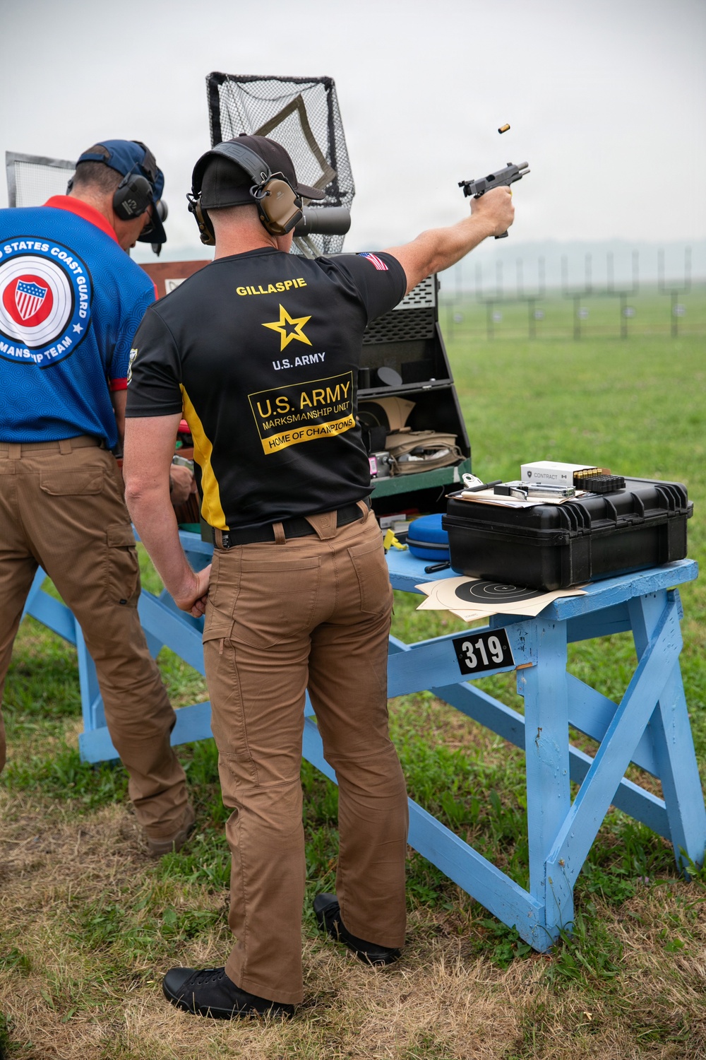 Two competitive shooters are at an outdoor pistol range, with the primary shooter identified as 'Gillaspie' from the U.S. Army Marksmanship Unit firing a semi-automatic pistol. A spent brass casing is visibly ejecting from the firearm mid-shot. The second individual is wearing a United States Coast Guard Marksmanship Team shirt and appears to be observing.