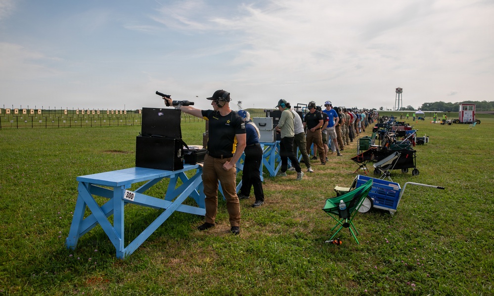 An outdoor pistol shooting competition is underway at a large range facility, with numerous competitors lined up along a row of blue shooting benches. The foreground shooter, wearing a black cap and ear protection, is actively firing a pistol downrange toward a series of paper targets. The setting appears to be a formal organized match, likely at a military or national-level shooting facility.