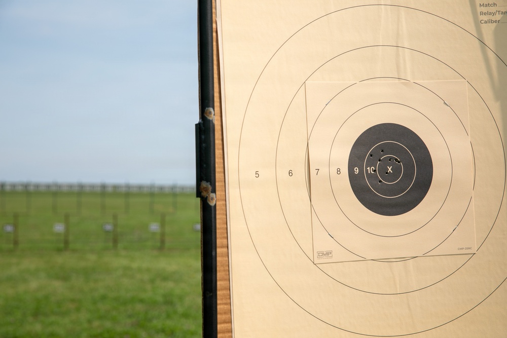 A CMP (Civilian Marksmanship Program) paper shooting target is mounted on a wooden board at an outdoor rifle range. The target shows several bullet holes concentrated in the high-scoring inner rings (X and 10 ring), indicating accurate shooting. In the background, a green field with additional target stands is visible under a partly cloudy sky.