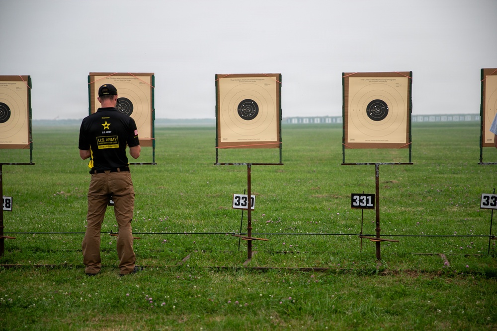 A U.S. Army competitive shooter is standing at an outdoor shooting range, examining or scoring targets on a grassy field under an overcast sky. The shooter is wearing a black U.S. Army Marksmanship Unit shirt and tan pants. Multiple numbered shooting targets (numbered 328-332 visible) are mounted on metal stands across the range.