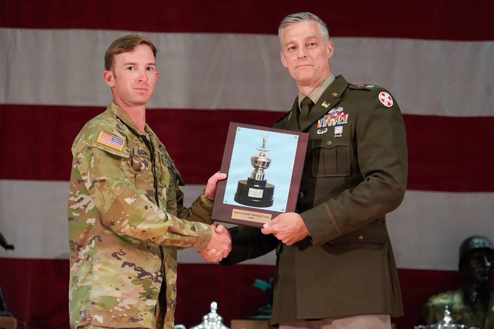 Two U.S. Army soldiers are participating in a formal award ceremony, shaking hands while jointly holding a plaque featuring a trophy image. The soldier on the left wears Army Combat Uniform (ACU/OCP pattern) with a name tape reading 'BUCHANAN,' while the officer on the right wears the Army Service Uniform (green) with multiple ribbons, rank insignia, and a red cross shoulder patch. An American flag serves as the backdrop.