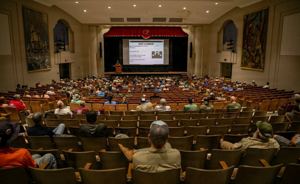 An auditorium or theater with wooden seats is partially filled with attendees watching a presentation on a projected screen. The screen displays a slide titled 'SIGHT ALIGNMENT' with instructional text and a diagram. The venue features decorative murals on the side walls and a stage with red curtains bearing the letters 'CP'.