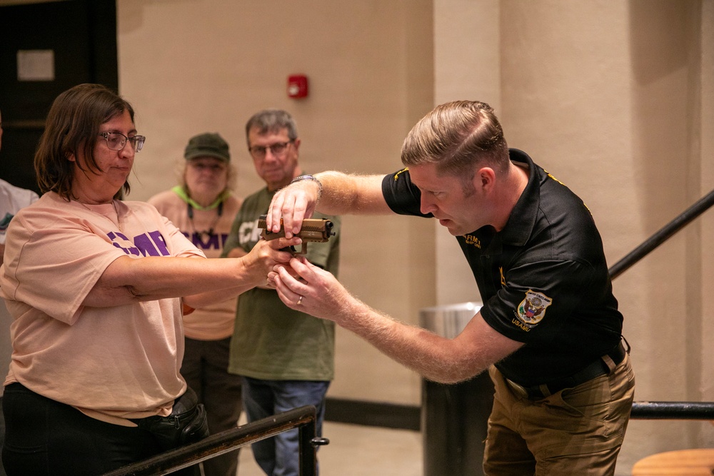 An instructor in a black uniform shirt with patches is demonstrating a firearm handling or disarmament technique with a civilian woman wearing a peach t-shirt with 'CMP' lettering. Two observers wearing green CMP lanyards/badges watch the demonstration from behind. The setting appears to be an indoor training or educational event, likely associated with the Civilian Marksmanship Program (CMP).