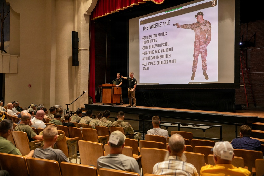 A military or law enforcement firearms training briefing is taking place in an auditorium-style theater. Two instructors on stage are presenting a slideshow about the 'One Handed Stance' pistol technique to an audience of predominantly uniformed military personnel. The projected slide displays bullet points describing proper stance mechanics alongside an image of a soldier demonstrating the technique.