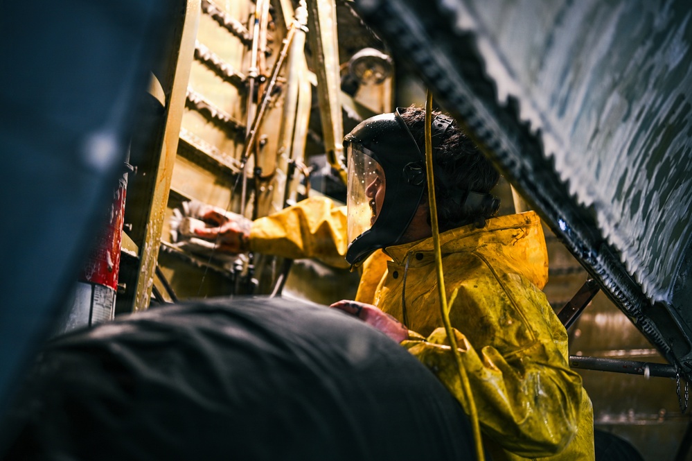 A worker wearing a yellow chemical/rain suit and a black protective face shield is performing maintenance or inspection work in a confined, industrial space. The environment appears wet and dimly lit, with pipes, metal structures, and machinery visible in the background. The setting suggests an industrial facility such as a ship hull, tunnel, or heavy industrial plant.