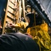 A worker wearing a yellow chemical/rain suit and a black protective face shield is performing maintenance or inspection work in a confined, industrial space. The environment appears wet and dimly lit, with pipes, metal structures, and machinery visible in the background. The setting suggests an industrial facility such as a ship hull, tunnel, or heavy industrial plant.