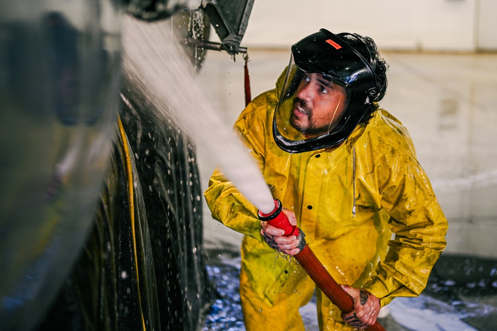 A worker in full protective gear is using a high-pressure hose to wash down a large dark-colored vehicle or aircraft in what appears to be an indoor facility. The individual is wearing a yellow rain suit and a black helmet with a face shield, suggesting a hazardous or high-pressure cleaning operation. Water and foam are visibly spraying against the surface being cleaned.
