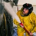 A worker in full protective gear is using a high-pressure hose to wash down a large dark-colored vehicle or aircraft in what appears to be an indoor facility. The individual is wearing a yellow rain suit and a black helmet with a face shield, suggesting a hazardous or high-pressure cleaning operation. Water and foam are visibly spraying against the surface being cleaned.