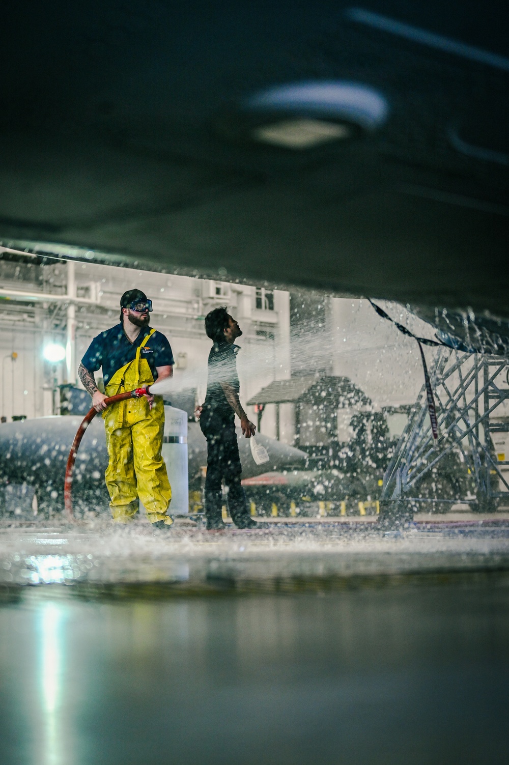 Two workers are engaged in a high-pressure washing operation beneath what appears to be a large aircraft or vessel in an industrial hangar facility. The primary worker in the foreground is wearing yellow protective rain gear and safety goggles while operating a red high-pressure hose, with water and spray visibly dispersing around them. A second worker stands nearby in dark clothing, appearing to observe or assist with the operation.