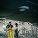 Two workers are engaged in a high-pressure washing operation beneath what appears to be a large aircraft or vessel in an industrial hangar facility. The primary worker in the foreground is wearing yellow protective rain gear and safety goggles while operating a red high-pressure hose, with water and spray visibly dispersing around them. A second worker stands nearby in dark clothing, appearing to observe or assist with the operation.
