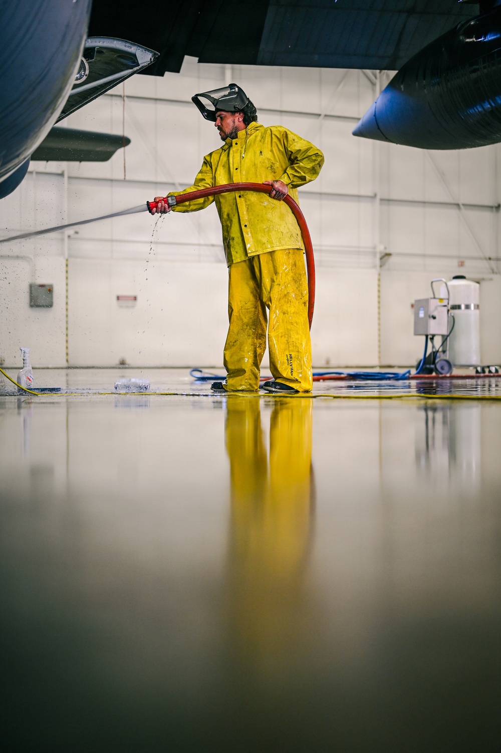 A worker in a yellow Helly Hansen rain suit and face shield is washing the interior floor of an aircraft hangar using a red hose. The wet, reflective epoxy floor creates a mirror-like reflection of the worker and the surrounding environment. Large aircraft components are visible on both sides of the frame, confirming the hangar setting.