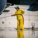 A worker in a yellow Helly Hansen rain suit and face shield is washing the interior floor of an aircraft hangar using a red hose. The wet, reflective epoxy floor creates a mirror-like reflection of the worker and the surrounding environment. Large aircraft components are visible on both sides of the frame, confirming the hangar setting.