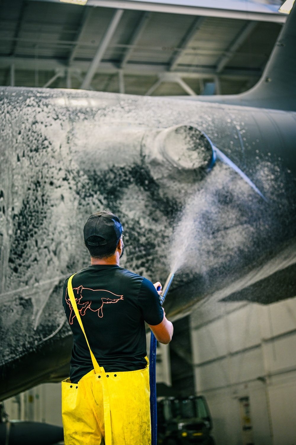 A worker in yellow waterproof overalls and a black t-shirt is pressure washing the underside or fuselage of a large dark-colored aircraft inside what appears to be a maintenance hangar. The surface being cleaned shows significant dirt, grime, and foam residue from the washing process. The setting is an industrial indoor facility with a metal ceiling structure visible overhead.