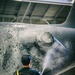 A worker in yellow waterproof overalls and a black t-shirt is pressure washing the underside or fuselage of a large dark-colored aircraft inside what appears to be a maintenance hangar. The surface being cleaned shows significant dirt, grime, and foam residue from the washing process. The setting is an industrial indoor facility with a metal ceiling structure visible overhead.